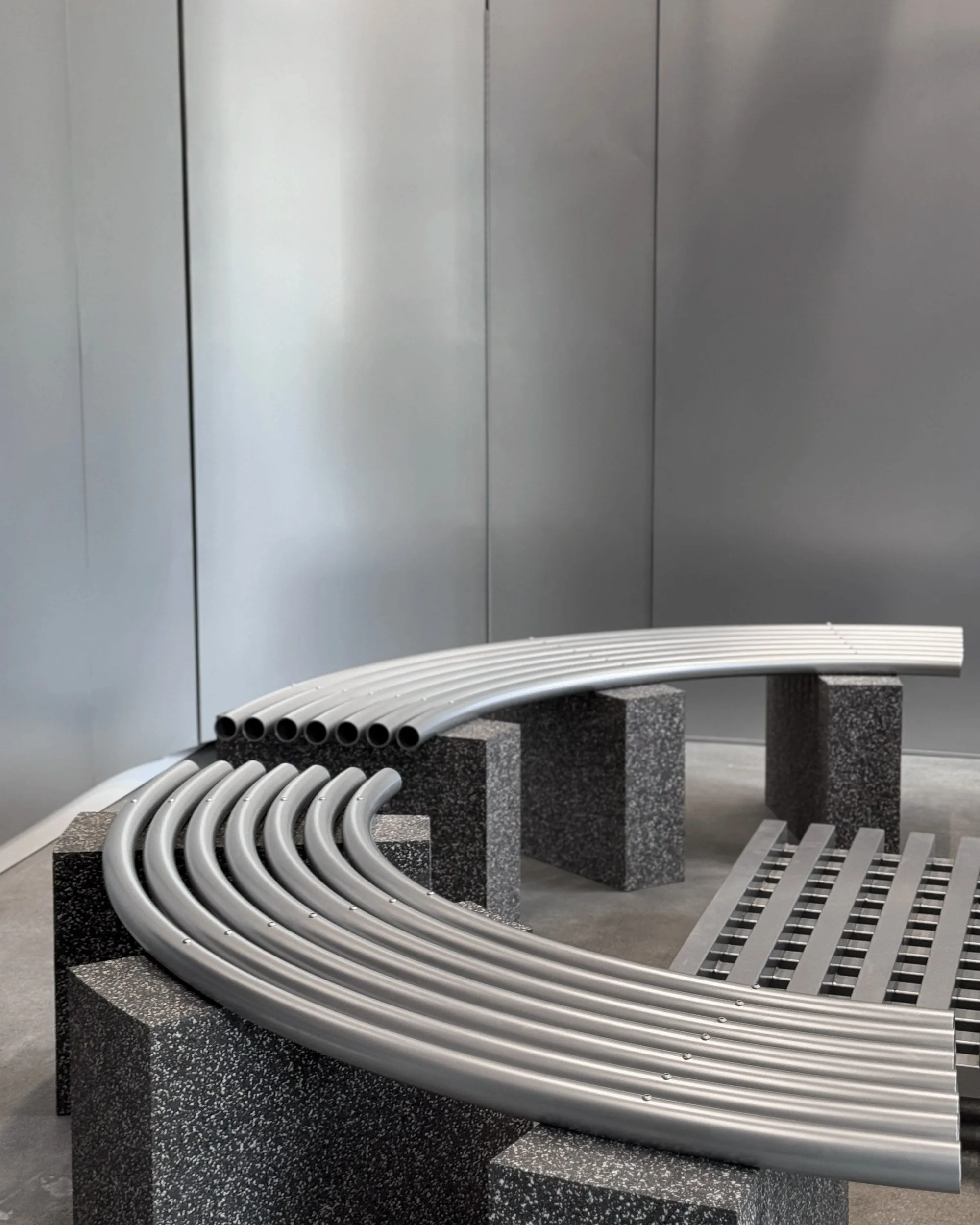 Curved aluminum tube bench resting on speckled stone blocks against a brushed metal wall.