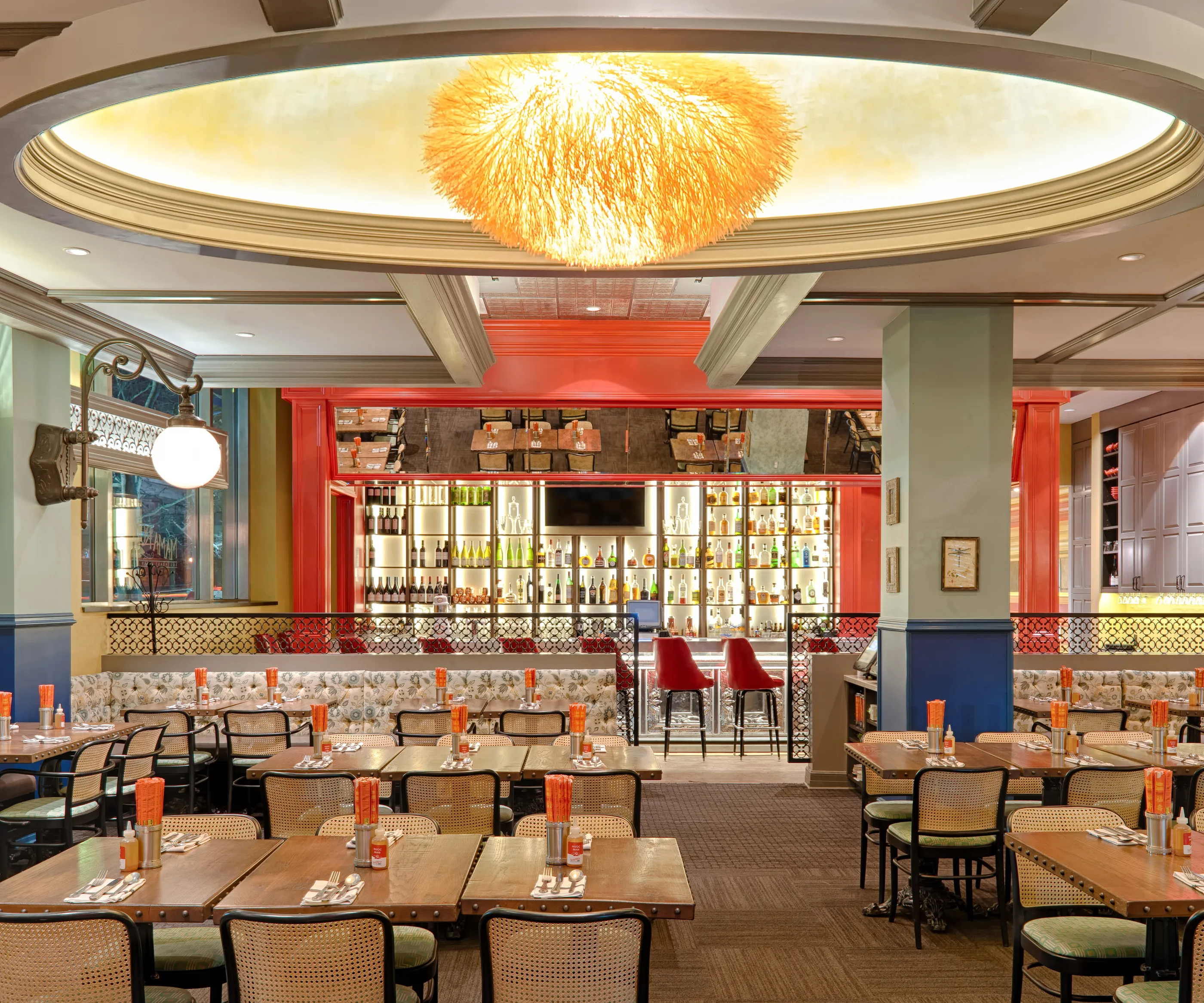 Restaurant dining room with timber tables, patterned banquettes, a red bar at the back, and a glowing oval ceiling with a chandelier.
