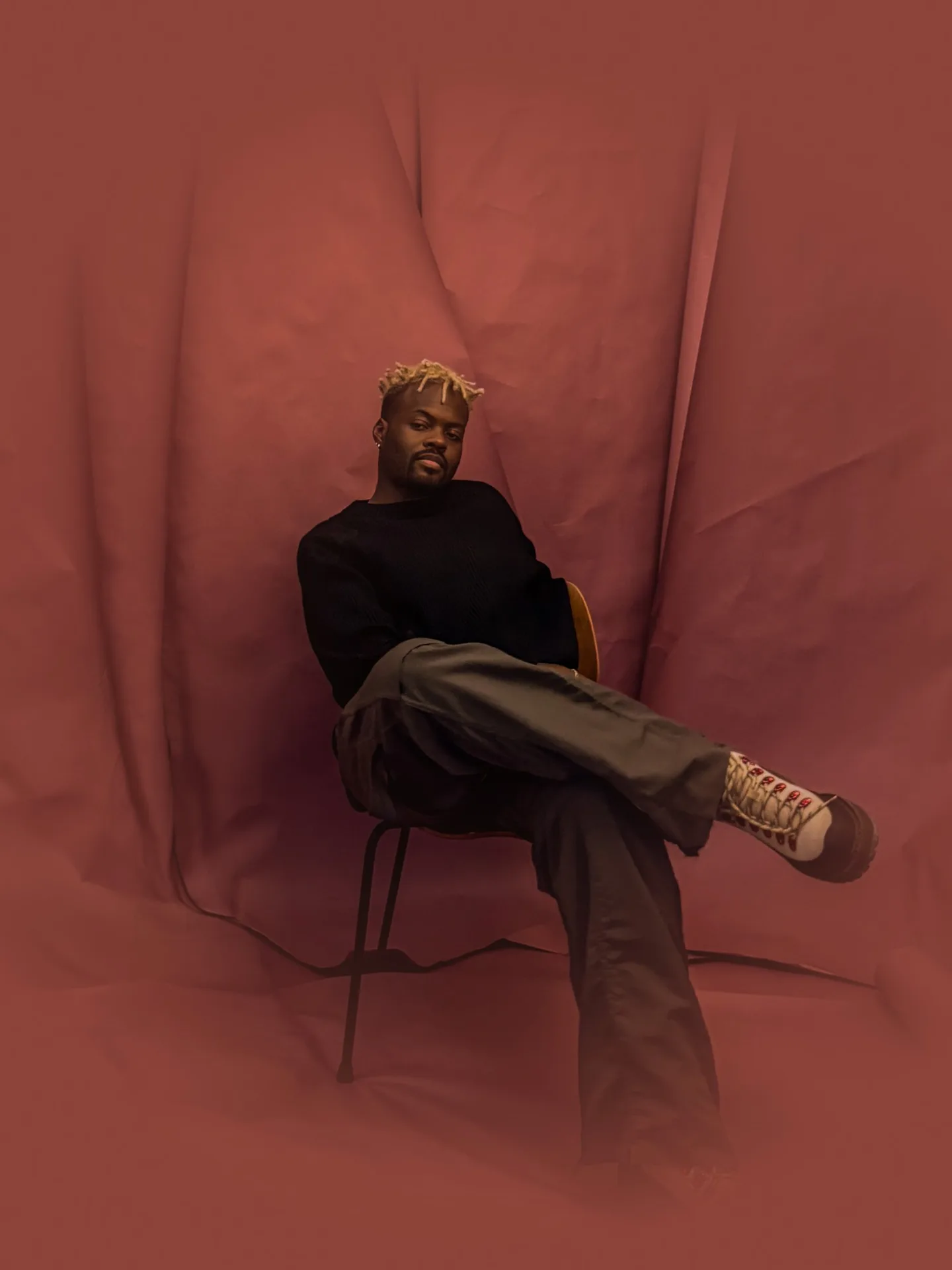 Jamel Williams seated on a wooden chair with legs crossed, in a black sweater and olive trousers, against a draped rust backdrop.