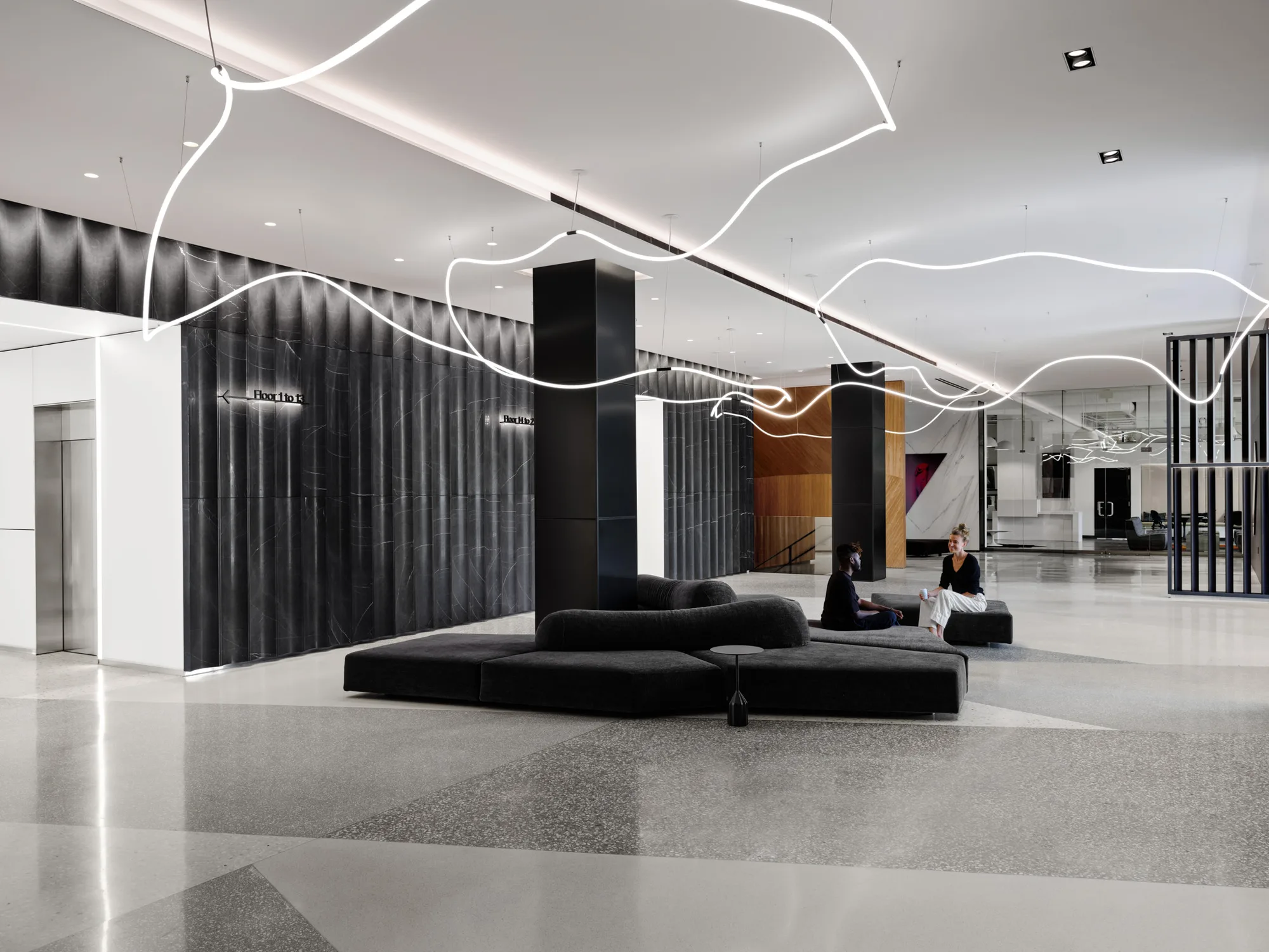 Wide lobby with dark panelled walls, looping white light lines on the ceiling, and low black seating with two people.