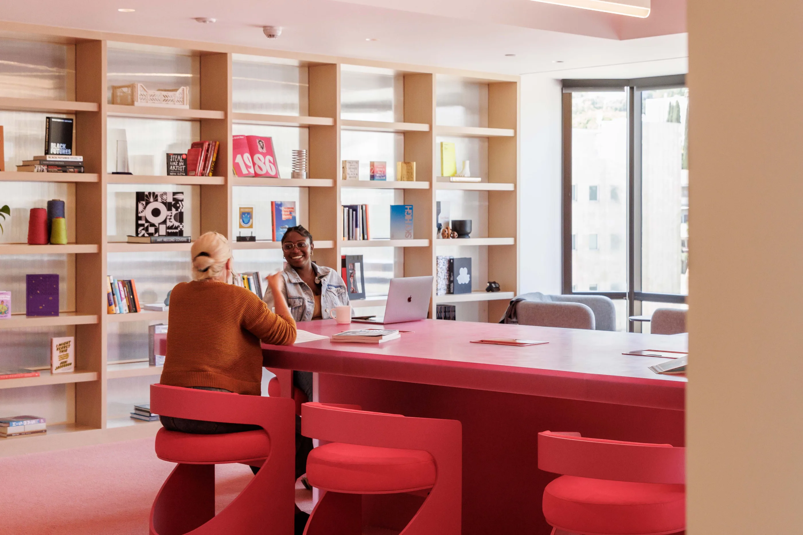 Library with pale wood shelves full of books and objects, a long pink table, and pink chairs.